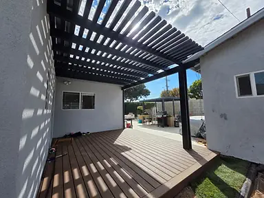 A charcoal-gray aluminum louvered pergola casts slatted shadows over a warm-brown composite deck attached to a white stucco home, bordered by a green hedge and low block wall under a partly cloudy sky.