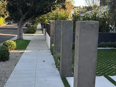 Row of smooth gray stucco pillars beside a concrete sidewalk and artificial-turf lawn.