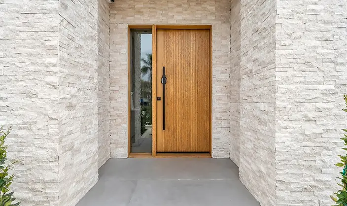 Close-up of new-construction entry at Valley Glen home: floor-to-ceiling stacked white stone surround framing a natural wood pivot door with black hardware.