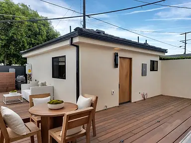 Spacious hardwood deck with teak dining set and lounge furniture beside a new-construction stucco ADU with wood-slat entry door, Valley Glen LA.