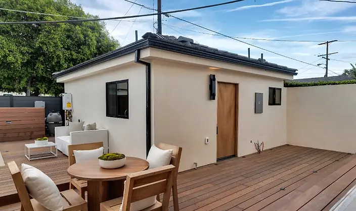 Spacious hardwood deck with teak dining set and lounge furniture beside a new-construction stucco ADU with wood-slat entry door, Valley Glen LA.