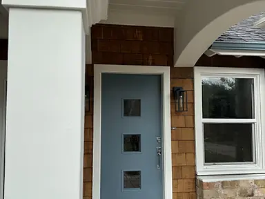 Freshly painted slate-blue modern front door with white frame and arch surround, white column, and cedar shingle siding on a residential home exterior.