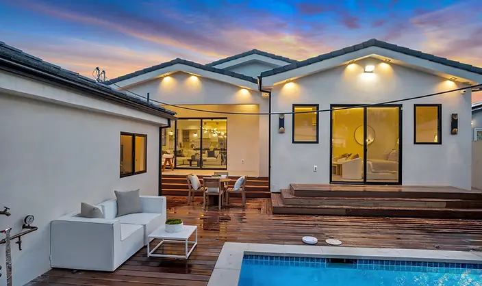 Modern white stucco home at dusk with a multi-level hardwood deck, outdoor furniture, string lights, and a rectangular pool with tile accents