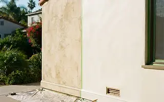 Editorial illustration: Stucco wall mid-repaint, half freshly coated and half faded and hairline-cracked, with masking and drop cloths