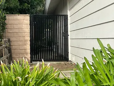 A dark charcoal-finished metal pedestrian gate with vertical flat-bar pickets is set between a stone-clad pillar and white stucco siding.