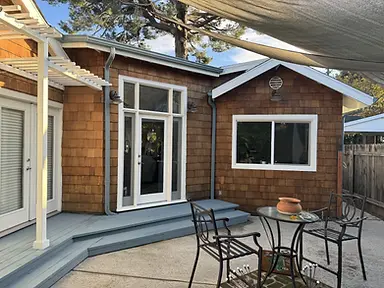 Backyard view of cedar shingle cottage with crisp white window trim and door frames, painted slate-blue deck and steps, and white pergola.