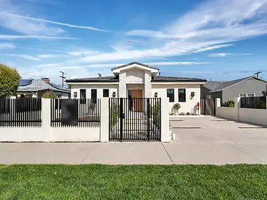 Street-facing view of new-construction home in Valley Glen with white stucco, black steel fence and gate, stone entry tower, and wide concrete driveway.