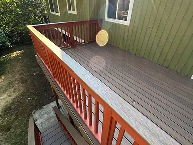 Completed raised composite deck beside a green-sided home, with redwood-stained railing, stairs, and a brown deck surface.