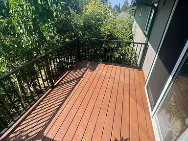 Completed composite balcony deck with a warm brown surface and black metal railing beside a gray home.