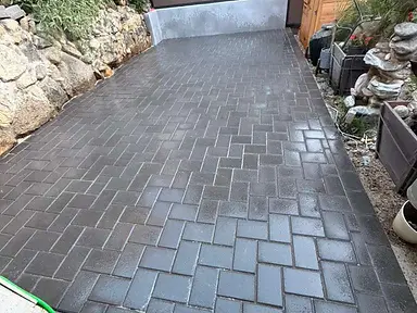 Charcoal grey concrete pavers laid in a herringbone pattern forming a side driveway between a stone retaining wall and a cedar fence