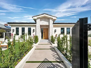View through open black steel gate of Valley Glen new home showcasing stacked-stone entry tower, wood pivot door, and freshly planted front garden.