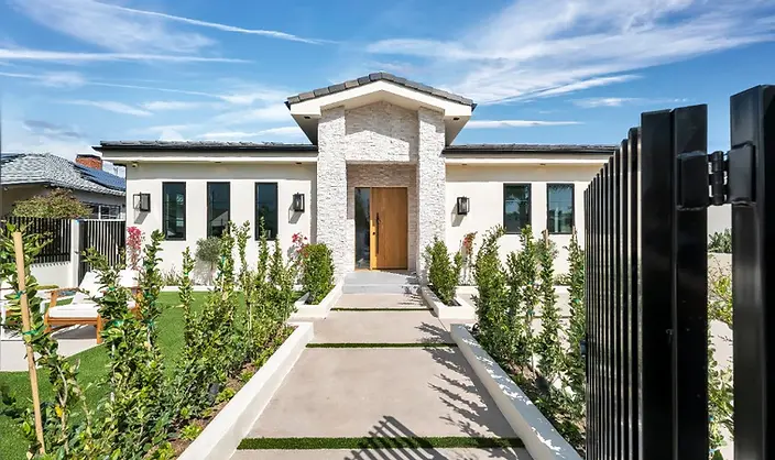 View through open black steel gate of Valley Glen new home showcasing stacked-stone entry tower, wood pivot door, and freshly planted front garden.