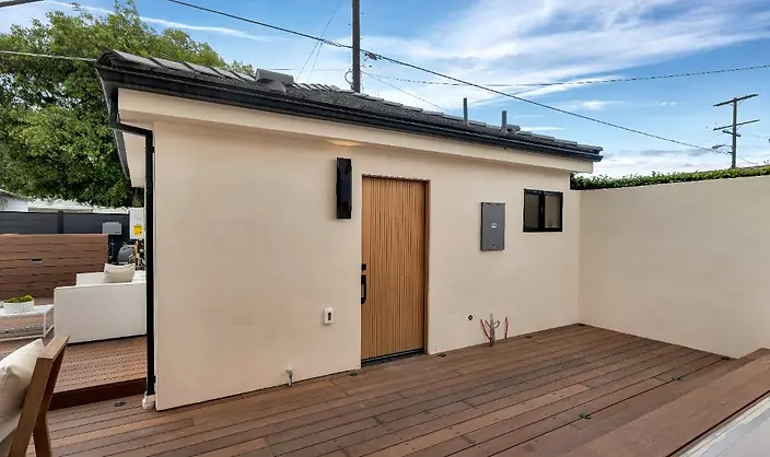 New-construction stucco ADU or garage structure in Valley Glen with warm ipe wood deck, slatted wood entry door, dark tile roof, and open-air patio area.