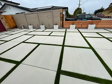Completed courtyard of large-format light concrete pavers with artificial-turf joints beside horizontal fencing and planters.
