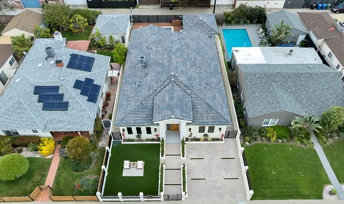 Drone aerial view of a completed new-construction home in Valley Glen, showing the concrete driveway, synthetic turf front yard, steel fence, and shingle roof.