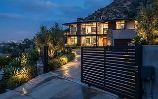 Editorial illustration: Automatic sliding aluminum driveway gate partially open across a sloped driveway at dusk with landscape lighting