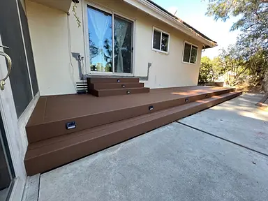 Completed low-profile brown composite deck with wide steps and integrated riser lights along a stucco home.
