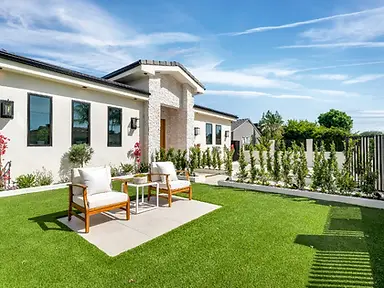 Rear yard of new-construction Valley Glen home with lush synthetic turf, concrete paver patio, teak lounge chairs, flowering shrubs, and stucco side elevation under blue sky.