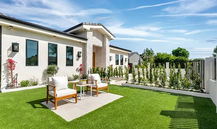 Rear yard of new-construction Valley Glen home with lush synthetic turf, concrete paver patio, teak lounge chairs, flowering shrubs, and stucco side elevation under blue sky.