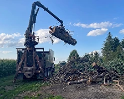 Grapple truck lifting a large log beside a cornfield under a clear sky