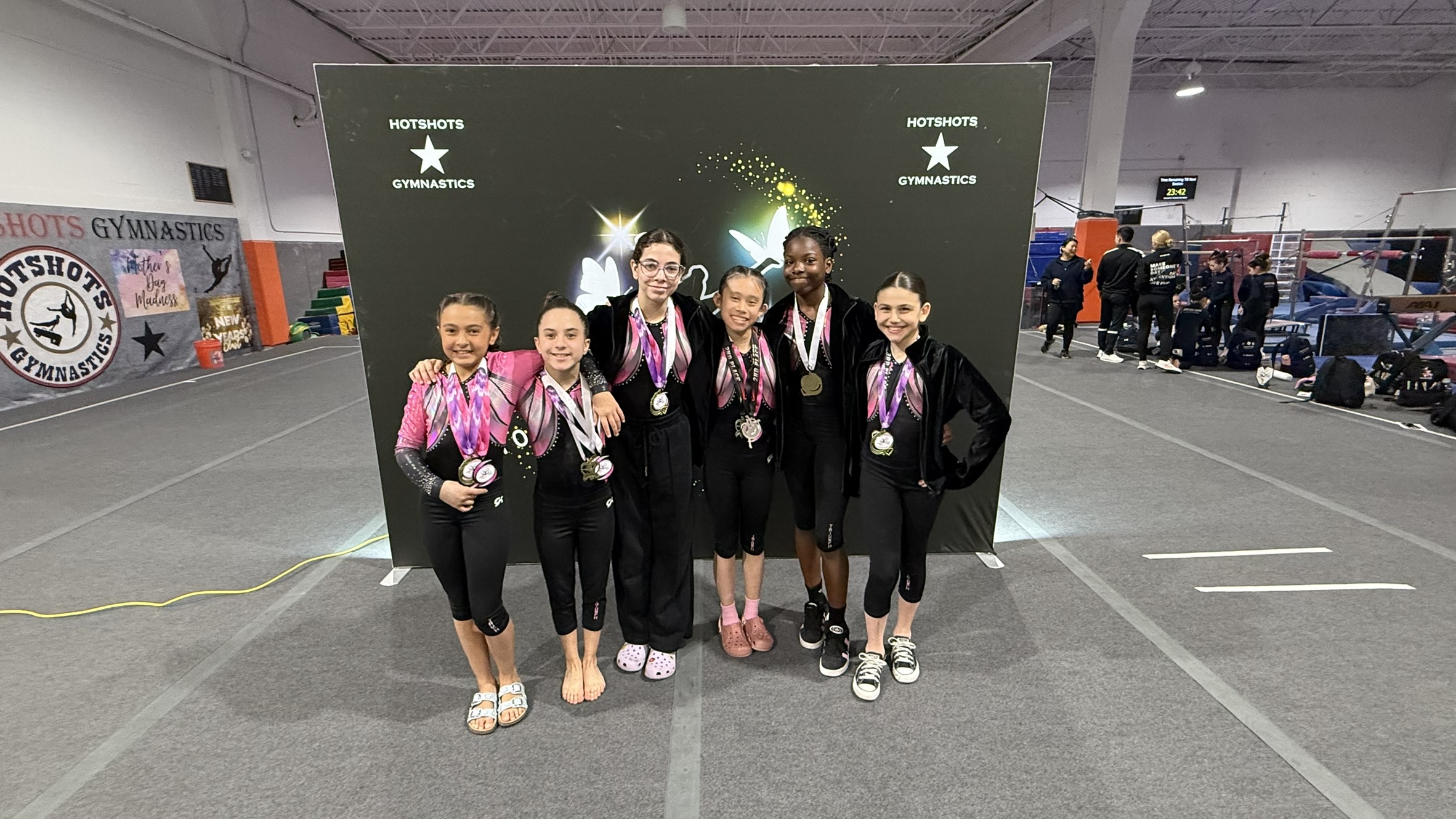 Triumph Gymnastics team gymnasts wearing medals and matching uniforms posing in front of a Hotshots Gymnastics backdrop