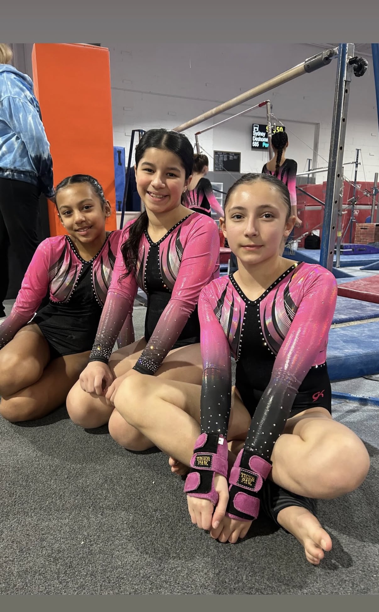 Three gymnasts in pink and black competition leotards sitting together