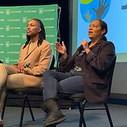 Two panelists sitting on stage at RootsCamp in front of a green RootsCamp backdrop, one in a tan blazer and one in a black blazer, both holding microphones