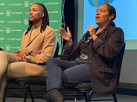 Two panelists sitting on stage at RootsCamp in front of a green RootsCamp backdrop, one in a tan blazer and one in a black blazer, both holding microphones