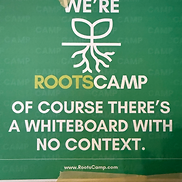 Green RootsCamp sign reading WE'RE ROOTSCAMP with the line 'OF COURSE THERE'S A WHITEBOARD WITH NO CONTEXT'