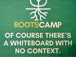 Green RootsCamp sign reading WE'RE ROOTSCAMP with the line 'OF COURSE THERE'S A WHITEBOARD WITH NO CONTEXT'