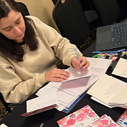 Person in a beige sweatshirt writing on documents at a black-clothed table during a RootsCamp session