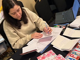 Person in a beige sweatshirt writing on documents at a black-clothed table during a RootsCamp session