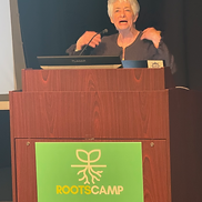 Woman with short white hair speaking at a wooden podium with a green RootsCamp sign during a session