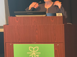 Woman with short white hair speaking at a wooden podium with a green RootsCamp sign during a session