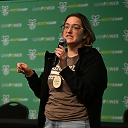 Melinda Frevert speaking into a microphone at a RootsCamp event, with a green step-and-repeat backdrop.