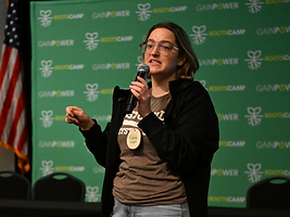 Melinda Frevert speaking into a microphone at a RootsCamp event, with a green step-and-repeat backdrop.
