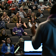 A packed RootsCamp auditorium during a keynote, viewed from behind the speaker's podium.