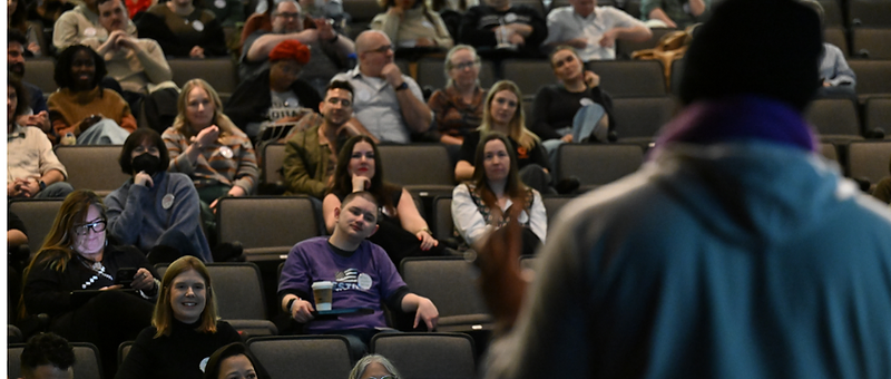 A packed RootsCamp auditorium during a keynote, viewed from behind the speaker's podium.