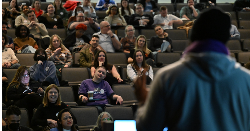 A packed RootsCamp auditorium during a keynote, viewed from behind the speaker's podium.