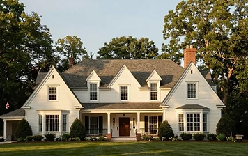 Symmetrical two-story white-sided estate home in Great Falls, VA with a steeply pitched charcoal-gray shingle roof, three gabled dormers, two red-brick chimneys, dark shutters, and a covered porch, framed by mature trees and a manicured lawn at golden hour