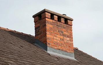 Close-up angled view of a traditional brick chimney with a concrete cap and dark step flashing protruding from a dark-brown shingled residential roof under a pale overcast sky
