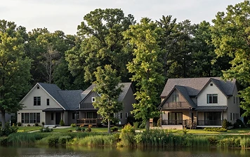 Two lakeside two-story homes in Reston, VA with dark-shingled roofs, light siding, and dark-trim balconies framed by tall reeds and dense mature deciduous trees in soft afternoon light