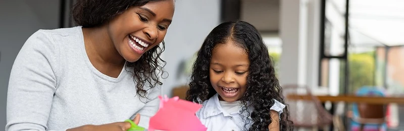 A woman and a young girl laughing together while cutting colorful construction paper at a table