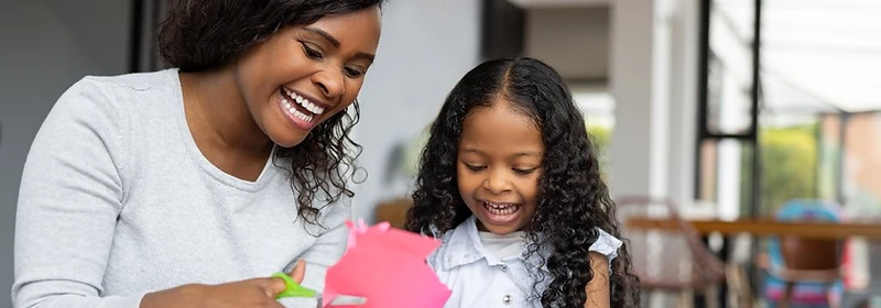 A woman and a young girl laughing together while cutting colorful construction paper at a table