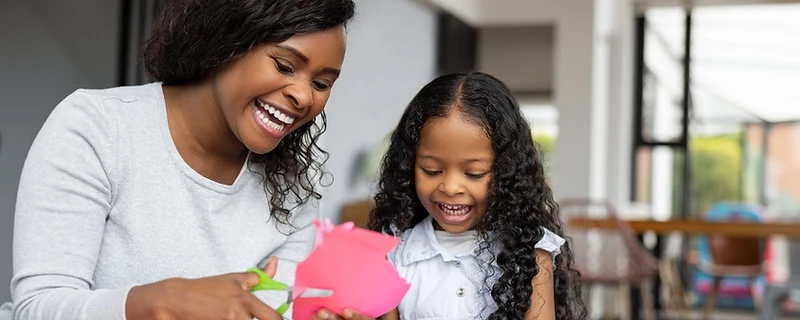 A woman and a young girl laughing together while cutting colorful construction paper at a table