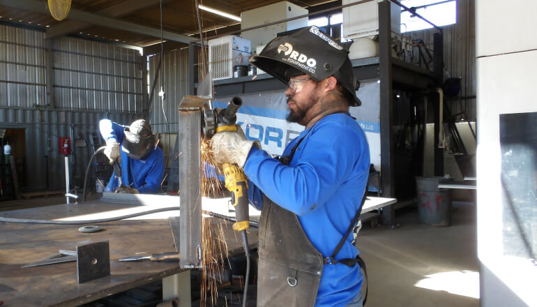 Metalworkers fabricating in the KorFab shop — grinder sparks flying on a steel beam