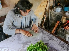 A person prepares dough on a lace-covered table beside fresh quelites and a traditional fogón with a small fire