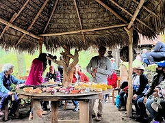 A campesino presents varieties of maize criollo on a circular table under a palapa to a gathered group of students and community members.