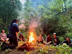 Participants gather around a campfire in a highland pine forest, holding an incense burner aloft during a field-based exchange of ritual and ecological knowledge.