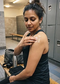 Adult woman in her early 30s applying a PACHEEZ patch on a gym locker room bench, smartphone style photo